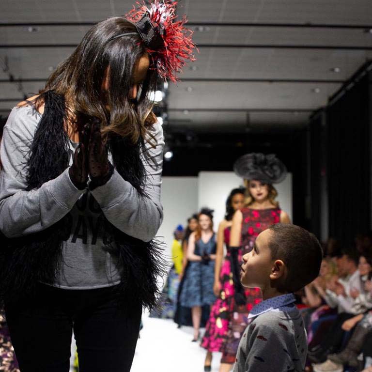 Fashion designer bowing in gratitude on runway with child in front and models in elegant dresses behind.