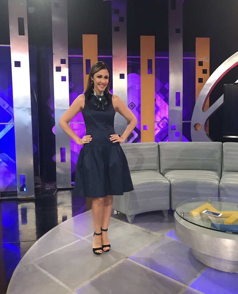 Mariasela Álvarez poses confidently on the set of her talk show, wearing a chic navy sleeveless dress with a flared skirt and black heels.