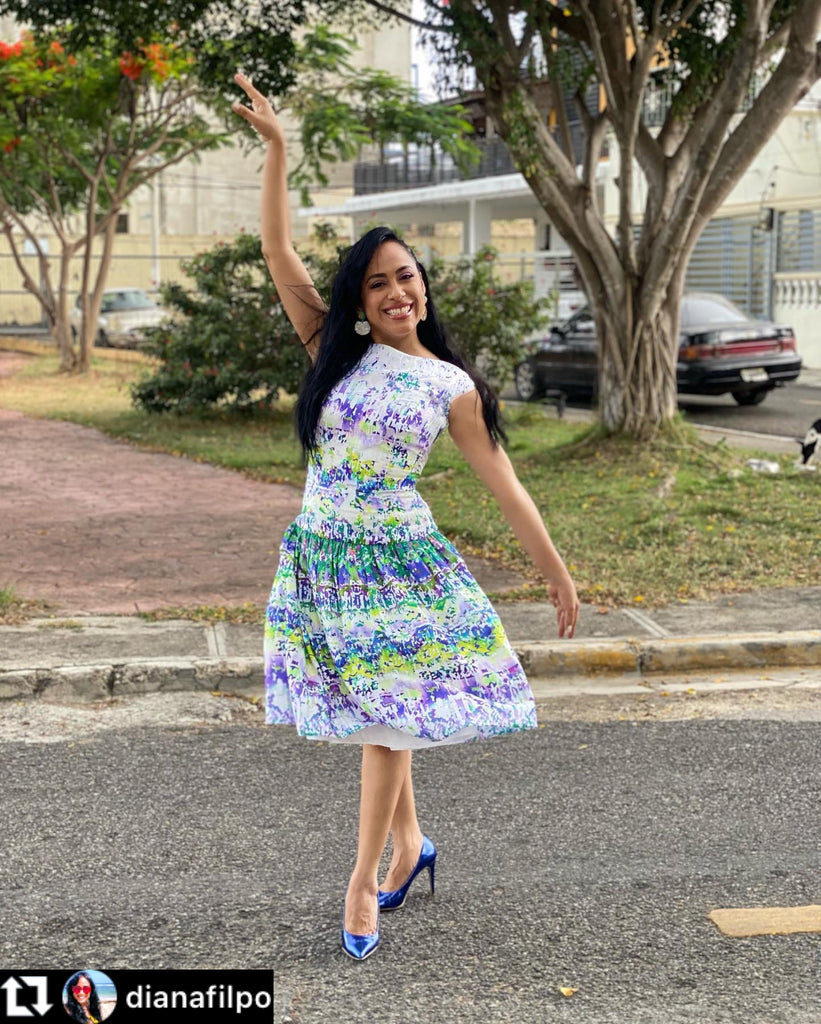 TV personality Diana Filpo wearing a sleeveless floral print dress in shades of purple, green, and white, paired with bright blue heels, posing gracefully outdoors on a tree-lined street.