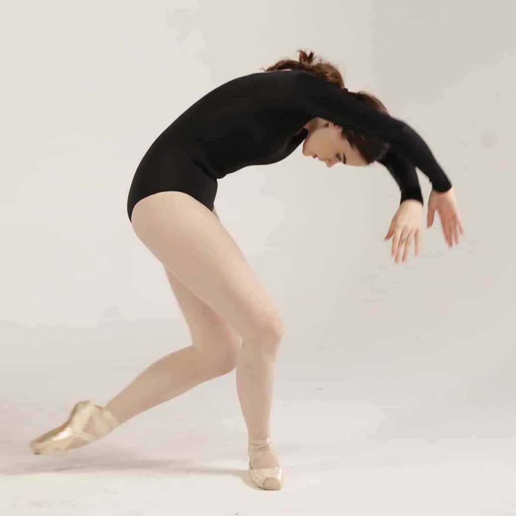 Ballet dancer in black leotard performing a graceful pose on pointe in a white studio setting.