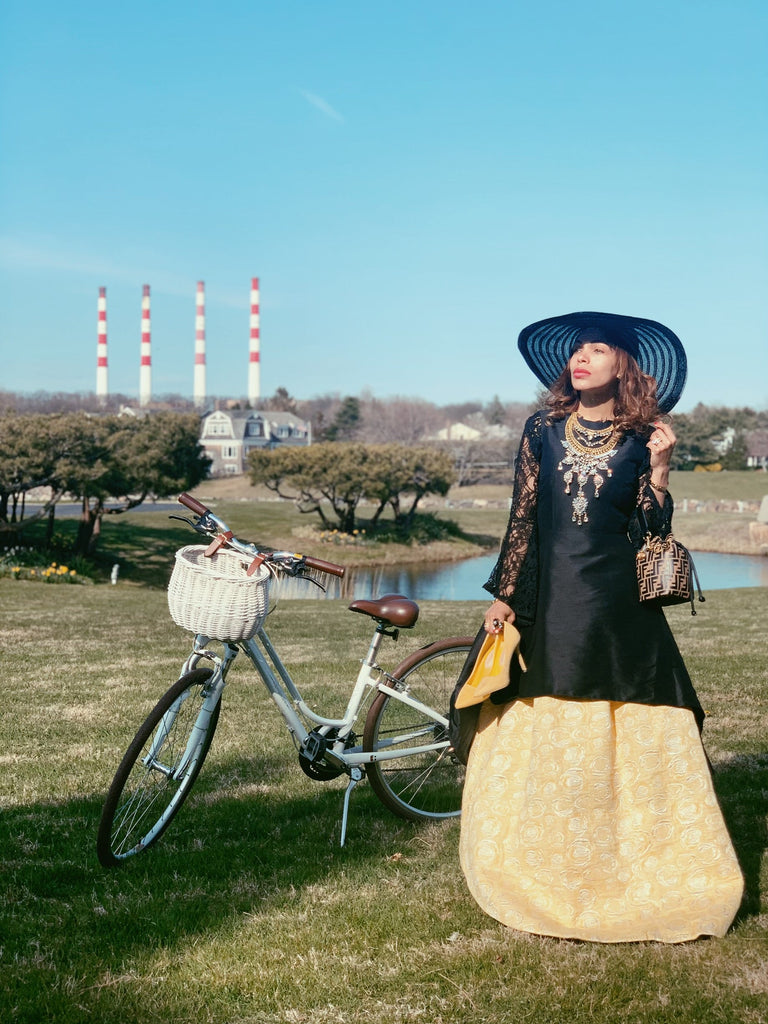 Angie Newlook posing outdoors beside a vintage-style bicycle, wearing a black and gold couture gown with lace sleeves and a wide-brimmed blue hat, showcasing Lalla Bee’s elegant custom design against a scenic grassy landscape.