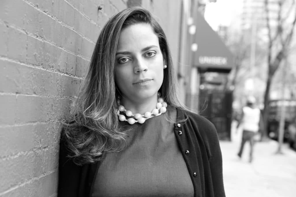 Melina Cortes-Nmili in a black-and-white portrait, wearing a statement necklace and leaning against a brick wall, embodying her roles as corporate leader, fashion designer, and mother.