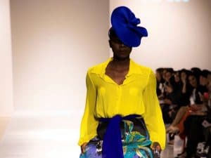 Model on the runway in a bright yellow blouse and bold floral skirt with blue sash, designed by Melina Cortes for Lalla Bee, featured in Emerson Today.