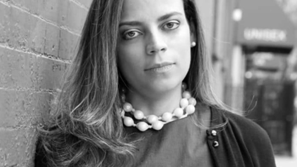 Black and white portrait of a woman wearing a pearl necklace and cardigan, standing against a brick wall.