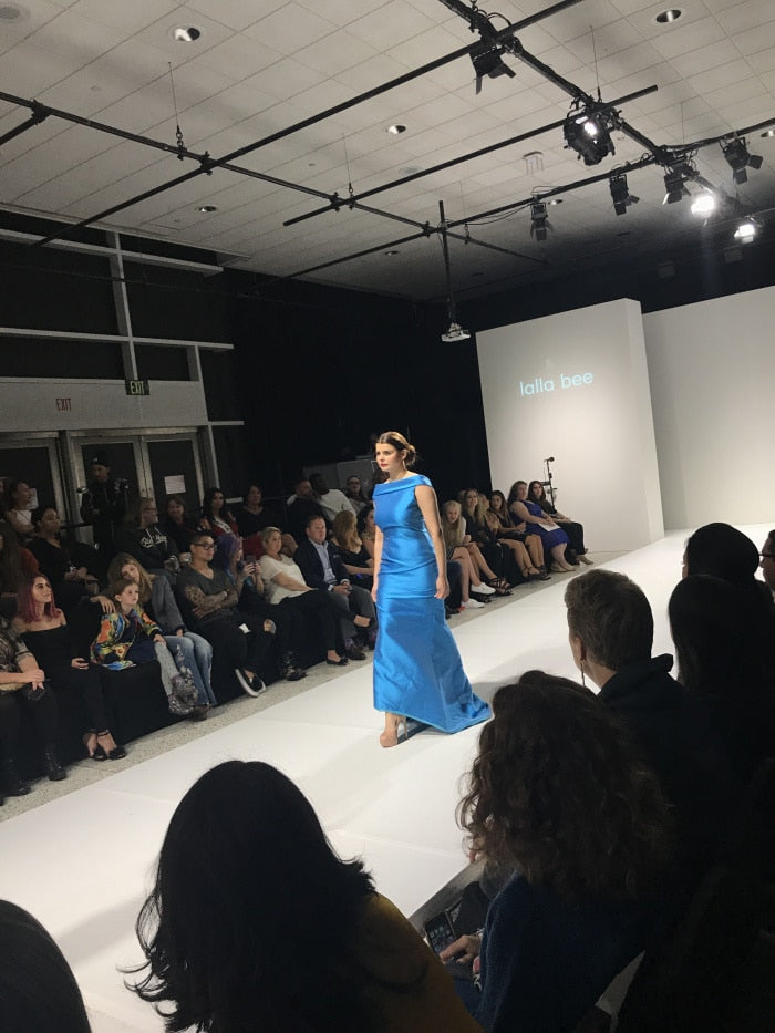 Model walking the runway in a sleek blue gown designed and made by Melina Cortes for Lalla Bee at StyleWeek Northeast Fall 2018, showcasing custom design, high end dresses, and luxury clothing for women by an independent luxury designer.