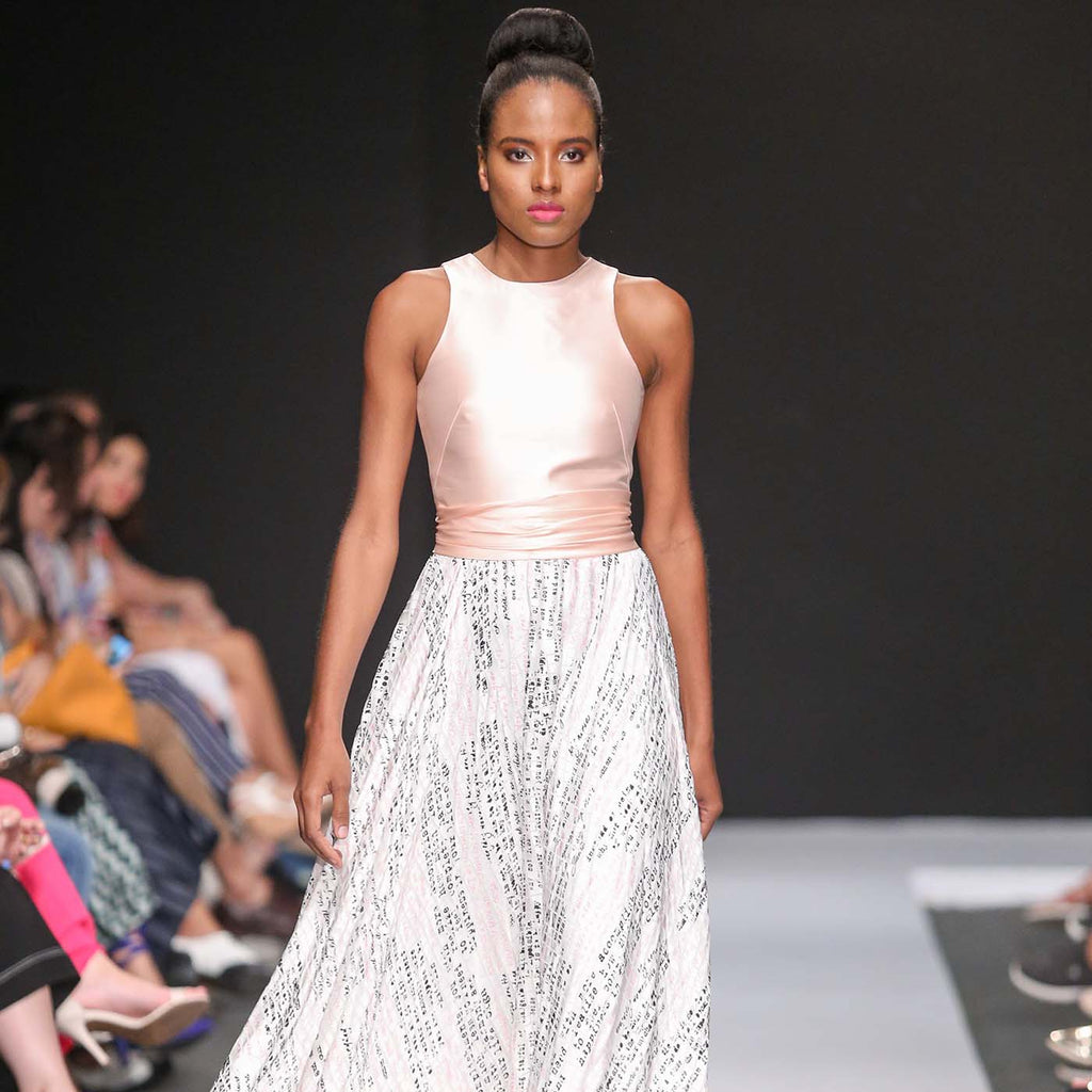 Model walking runway in sleeveless blush pink top and flowing white skirt with printed text design.