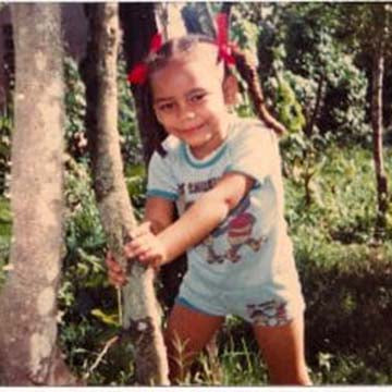 Vintage photo of a young girl with braids and red ribbons smiling while holding onto a tree in a green outdoor setting.