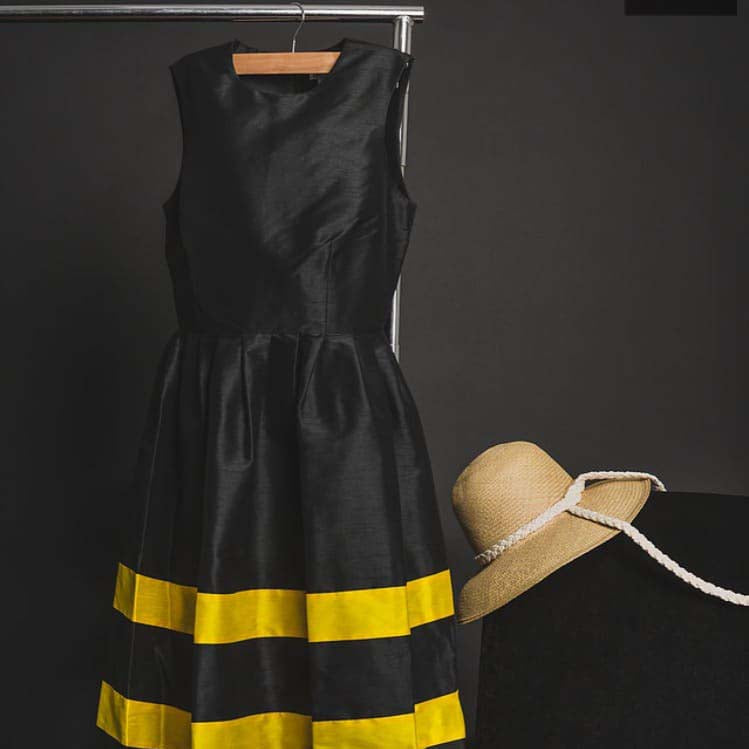 Black sleeveless dress with yellow stripes on hanger next to straw hat.