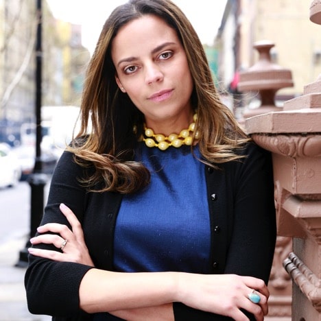 Woman in a blue dress with a statement necklace and black cardigan standing outdoors with arms crossed.