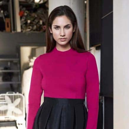 Woman in a fuchsia long-sleeve top with a black pleated skirt standing indoors.