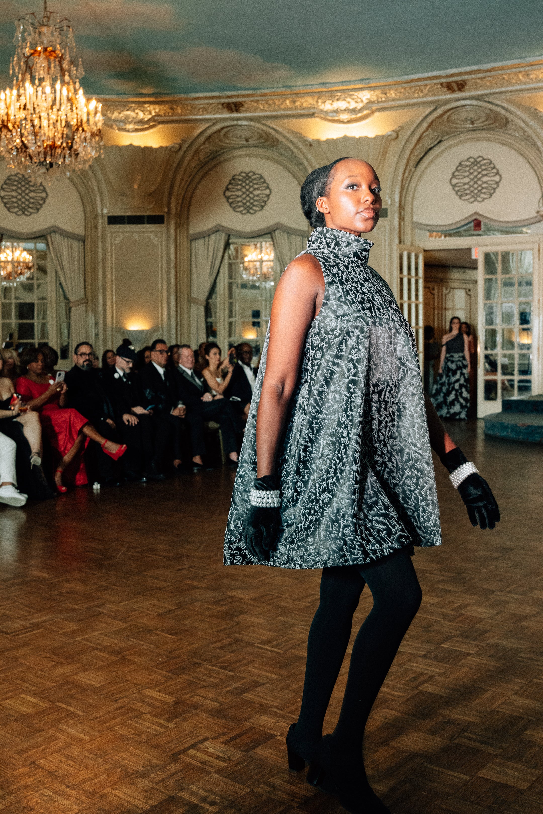 Model wearing the Lalla Bee Betty, a sleeveless high-neck black and silver patterned swing dress with an A-line silhouette, paired with black tights and gloves, perfect for luxury gatherings.